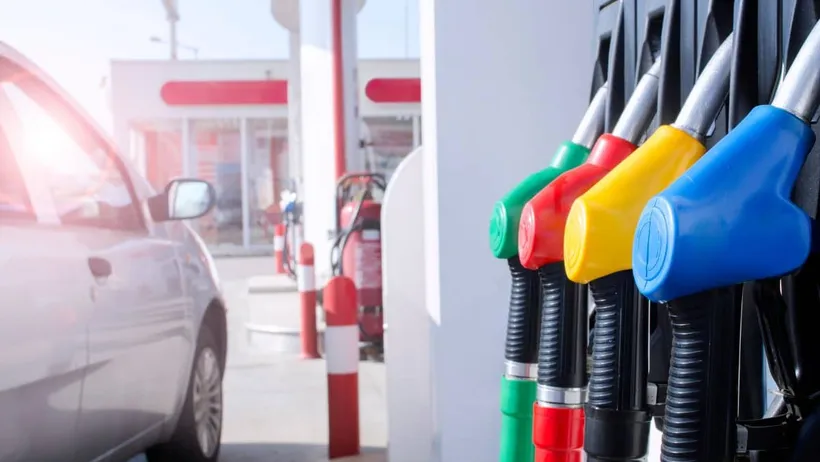 imagem de um carro popular de cor cinza ao lado de uma bomba de combustíveis em um posto de gasolina