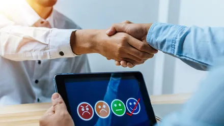 Cliente e revendor apertando as mãos sobre um tablet com figuras simulando as expressões de satisfação humana para simbolizar a retenção de clientes.