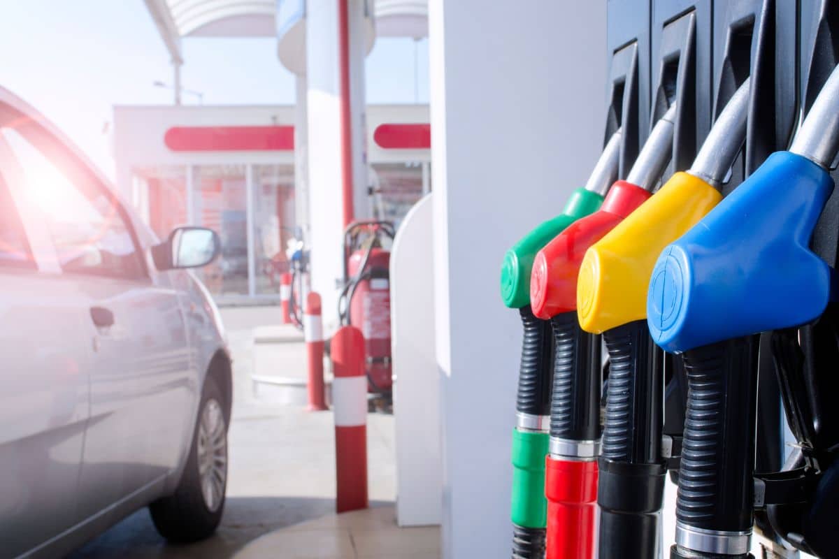 imagem de um carro popular de cor cinza ao lado de uma bomba de combustíveis em um posto de gasolina